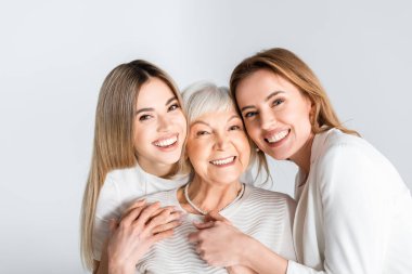 three generation of positive women smiling while looking at camera and hugging isolated on grey clipart