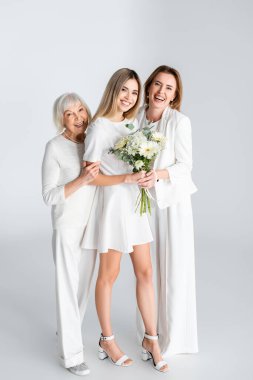 Anne ve büyükannenin yanında çiçeklerle gülümseyen mutlu genç bir kadın.