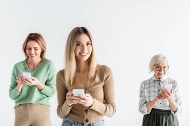 Annesinin yanında akıllı telefonuyla duran mutlu genç bir kadın ve bulanık arka planda beyaz bir kadın.