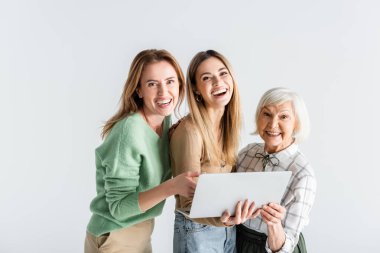 Bilgisayarın yakınındaki kameraya bakan üç nesil mutlu kadın beyaz bilgisayardan izole edilmiş.