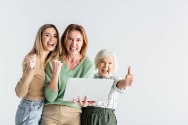 Kameraya bakan, başparmağını kaldıran ve dizüstü bilgisayarını beyaz ekranda tutan üç neslin mutlu kadını.