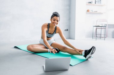 Gülümseyen Afro-Amerikan kadın spor giyimli evde dizüstü bilgisayarın yanında spor minderinde oturuyor.