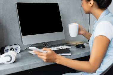 Beyaz ekranlı, bulanık önplanı olan, elinde akıllı telefon ve çay fincanı tutan Afrikalı Amerikalı serbest yazar görüntüsü.