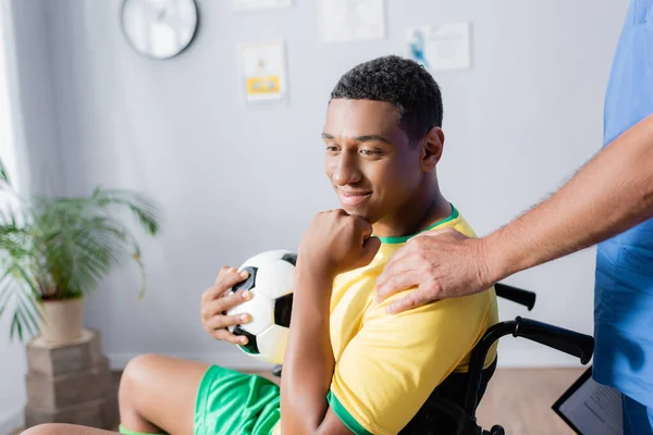 Lekarz Dotykający Ramienia Uśmiechniętego Afrykańskiego Sportowca Wózku Inwalidzkim Trzymającego Piłkę — Zdjęcie stockowe