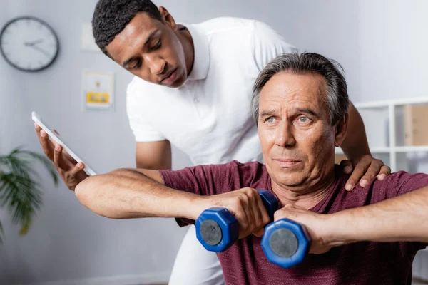 Fisioterapeuta Segurando Tablet Digital Olhando Para Homem Meia Idade Exercitando — Fotografia de Stock