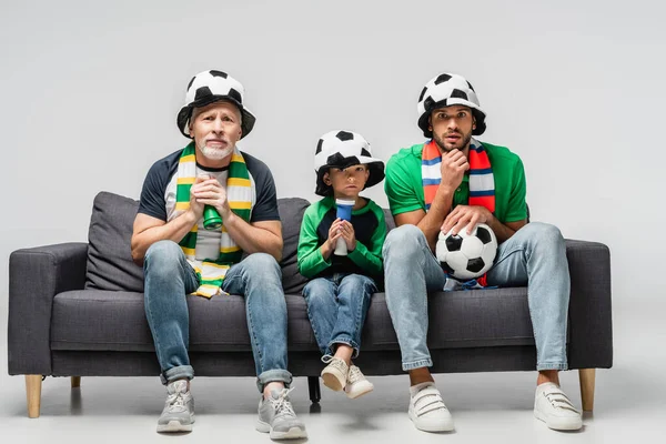 worried men and boy in fan hats watching football championship while sitting on sofa on grey
