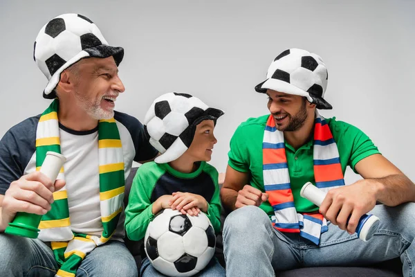 happy men in fan hats holding horns near boy with soccer ball isolated on grey