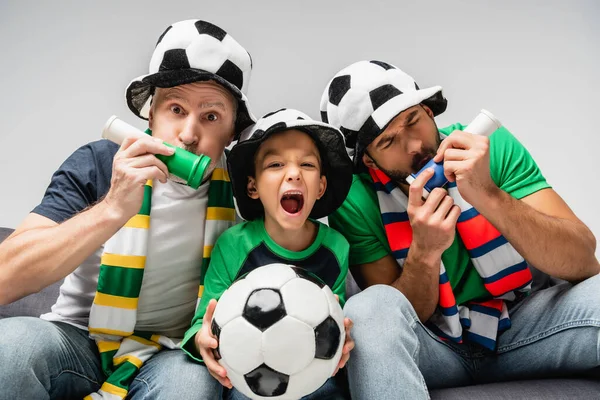 men in fan hats blowing in horns near excited boy screaming while holding soccer ball isolated on grey