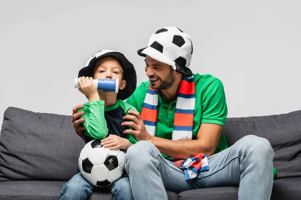 戴着足球迷帽的快乐男人拥抱着儿子 吹着被灰色隔离的号角 — 图库照片