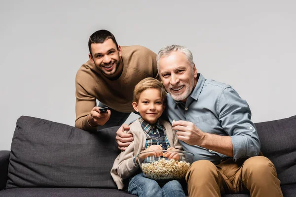 快乐的男人在父亲和儿子身边牵着遥控器看电视 吃着灰色的爆米花 — 图库照片