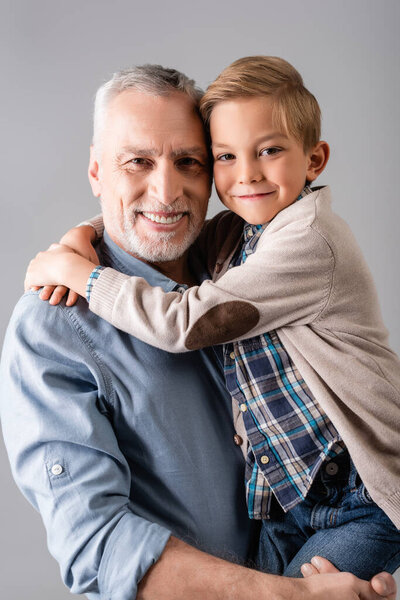 веселый мужчина улыбается в камеру, держа счастливого внука, обнимающего его изолированным на сером