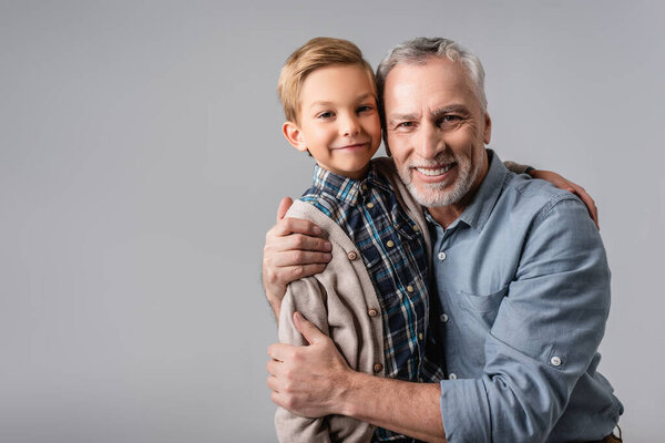 счастливый зрелый мужчина обнимает улыбающегося внука, глядя на камеру, изолированную на сером