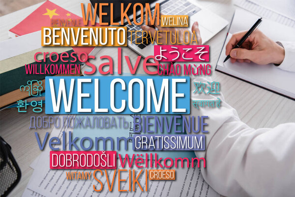 partial view of translator writing in notebook near laptop and dictionaries, words in different languages illustration. Translation: "welcome"
