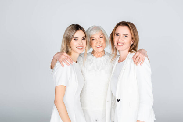 три поколения веселых женщин улыбаются, глядя в камеру, и обнимаются на сером фоне