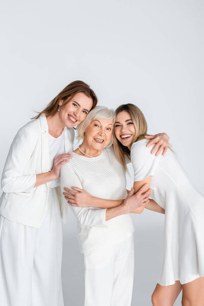 three generation of pleased women smiling while looking at camera and hugging isolated on grey