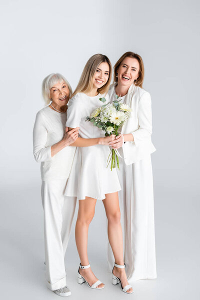 full length of happy young woman smiling while holding flowers near mother and granny on grey
