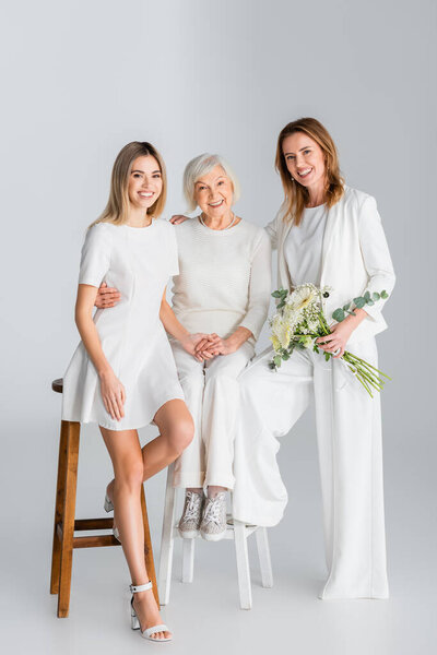 полная длина счастливой женщины, которая держит цветы и улыбается, сидя на стуле с дочерью и матерью на сером