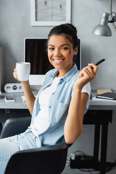 快乐的非洲裔美国自由职业者 坐在电脑前 手里拿着一杯茶和一支笔 — 图库照片