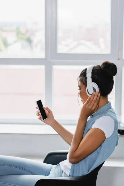 自宅の窓の近くに座っている間にスマートフォンを持っているワイヤレスヘッドフォンの若いアフリカ系アメリカ人女性 — ストック写真