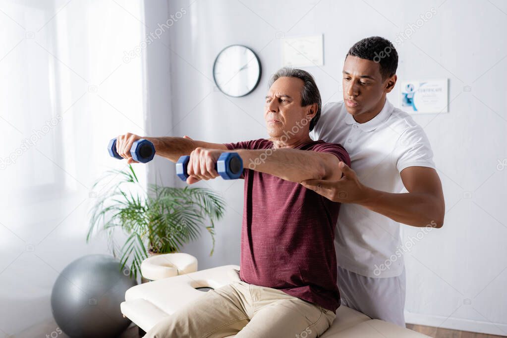 middle aged man exercising with dumbbells on massage table near african american physiotherapist 
