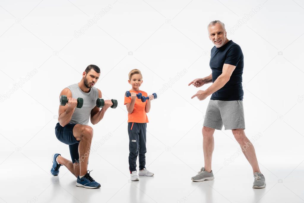 happy man pointing with fingers at father and son exercising with dumbbells on white