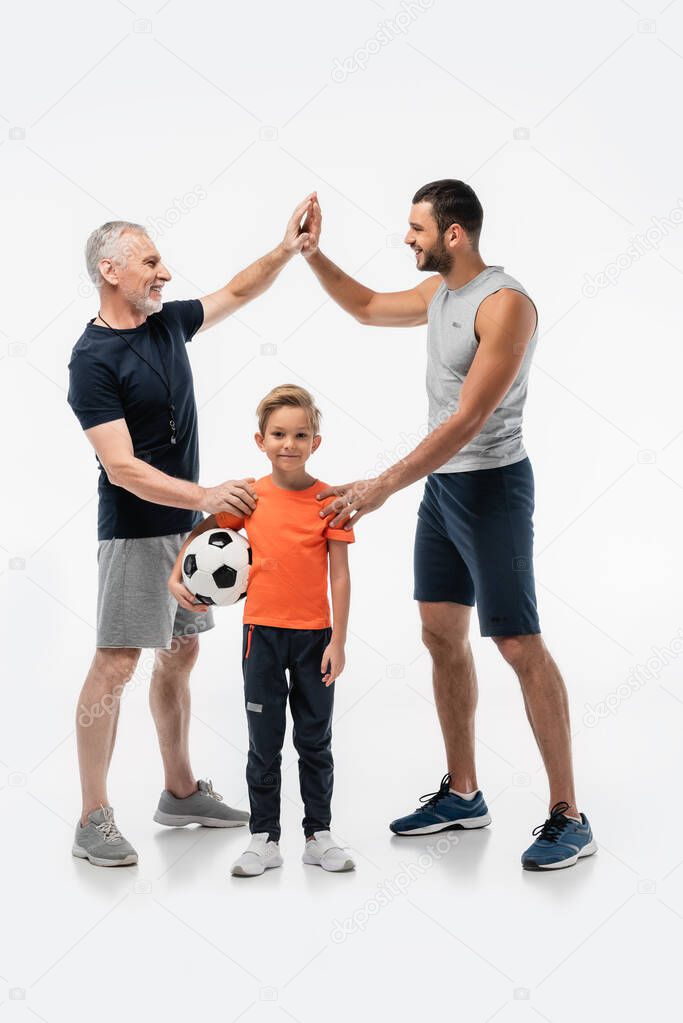 happy boy holding soccer ball and smiling at camera near dad and grandfather giving high five on white