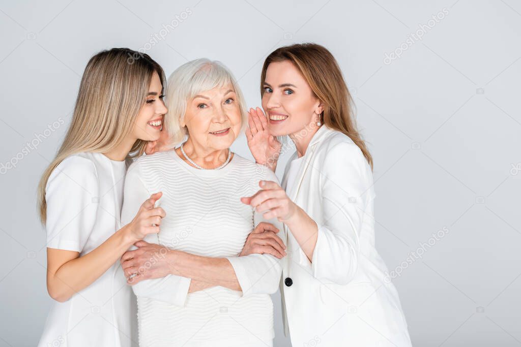 happy woman whispering in ear of senior mother near young daughter isolated on grey