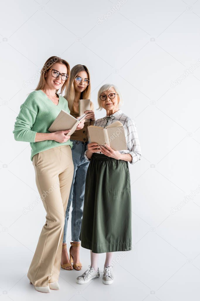 full length of three generation of smiling women in glasses holding books on white