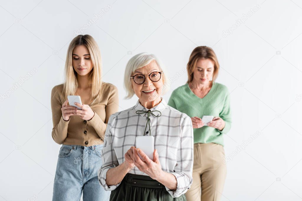 cheerful senior woman in glasses holding smartphone near daughter and granddaughter on blurred background