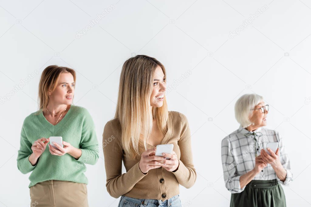 cheerful young woman standing with smartphone near mother and granny on blurred background isolated on white