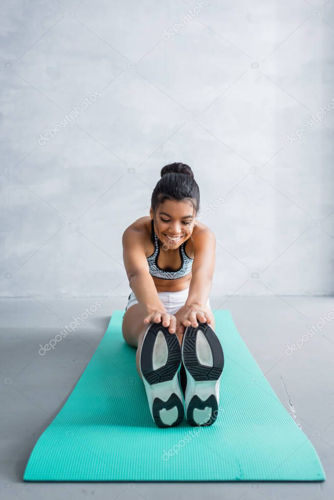 smiling african american sportswoman doing seated forward bend exercise at home