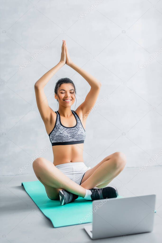happy african american woman sitting in lotus pose with praying hands on fitness mat near laptop