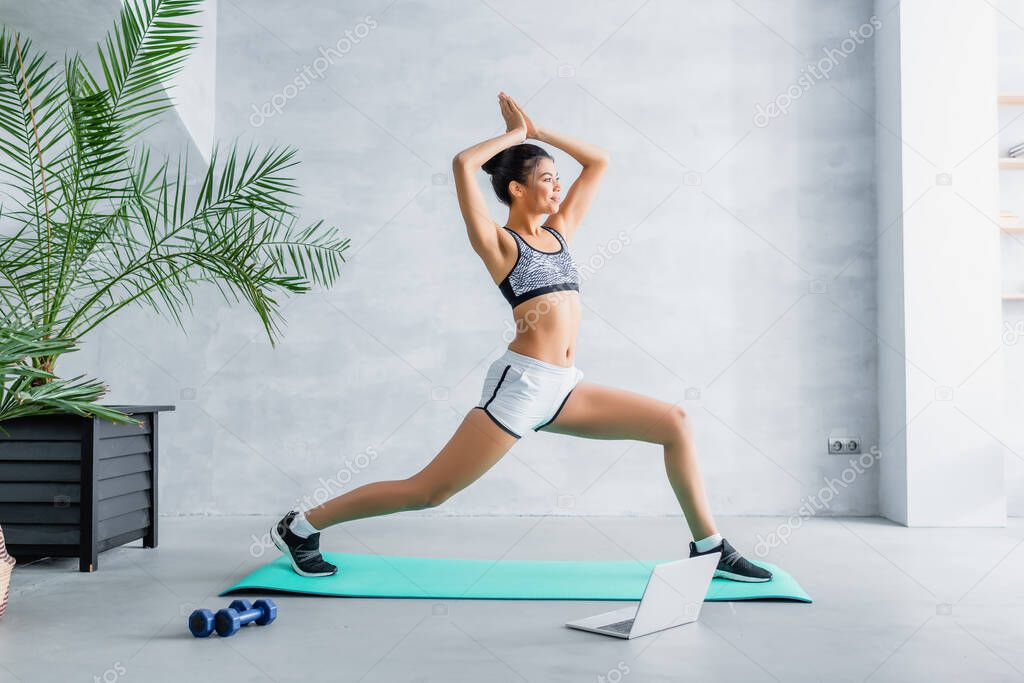 young african american sportswoman exercising in warrior pose near laptop and dumbbells at home
