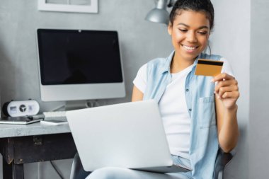 Bilgisayarı ve kredi kartı olan mutlu bir Afrikalı Amerikalı kadın. Geçmişi bulanık, boş ekranlı bir bilgisayar monitörü yanında.
