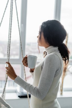 Genç Afrikalı Amerikalı kadın elinde bir fincan sıcak çay tutarken pencereden bakıyor. 