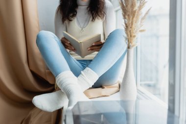 cropped view of african american woman reading book while sitting on windowsill with crossed legs clipart