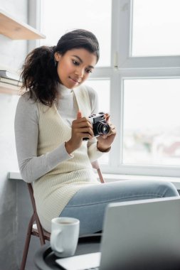 Genç Afro-Amerikalı kadın elinde eski model fotoğraf makinesi tutarken ön planda bulanık dizüstü bilgisayara bakıyor.