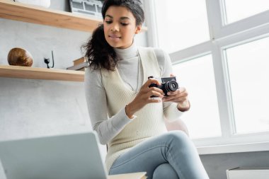 Genç Afro-Amerikalı kadın elinde eski model fotoğraf makinesi tutarken laptopun yanında bulanık ön planda oturuyor.