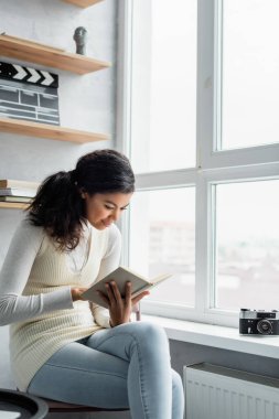 young african american woman reading book while sitting near vintage photo camera on windowsill clipart