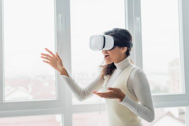 VR kulaklıklı Afro-Amerikalı kadın evdeki pencerenin yanında el kol hareketi yapıyor.