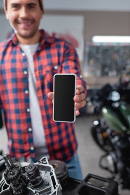 gülümseyen tamirci motorsiklet yedek parçalarının yanında boş ekranlı akıllı telefon gösteriyor, bulanık arkaplan