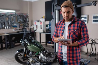 young mechanic in beanie and plaid shirt using digital tablet near spare parts and motorcycle on blurred background clipart