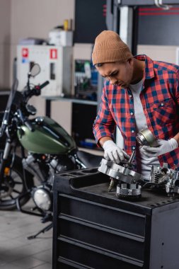 young technician in plaid shirt and beanie checking parts of disassembled gearbox near motorbike on blurred background clipart
