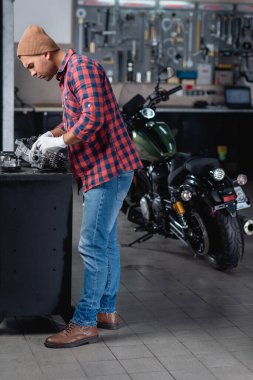 Ekose gömlekli ve kot pantolonlu tamircinin tam boy görünümü atölyede motosikletin yanında sökülmüş vites kutusunu kontrol ediyor