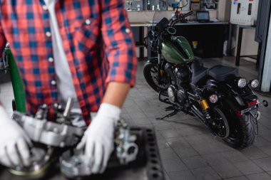 selective focus of motorcycle near mechanic and disassembled gearbox on blurred foreground clipart