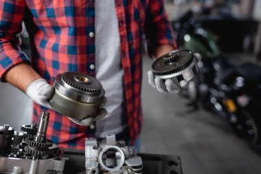 Ekose gömlekli ve eldivenli tamircinin kısmi görüntüsü motorsiklet şanzımanının dişlileri, bulanık arka plan