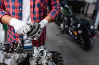 cropped view of mechanic in plaid shirt and gloves holding gearwheel near disassembled motorbike transmission clipart