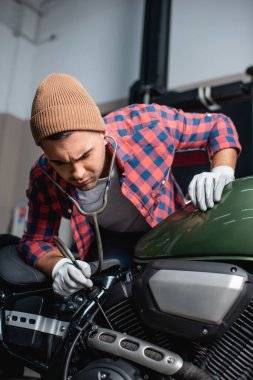Ekose gömlekli ve şapkalı bir tamirci steteskoplu motosiklet motoru tanısı yapıyor.