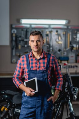 Tulumlu tamirci, arka planda bulanık motosikletin yanındaki kameraya bakıyor.