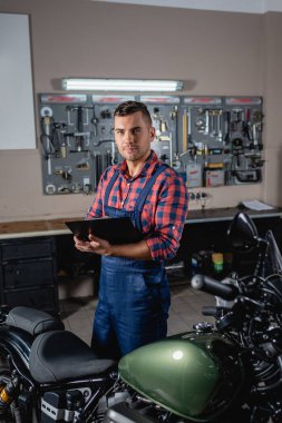 Genç teknisyen elinde panoyla motosikletin yanında dururken kameraya bakıyor.